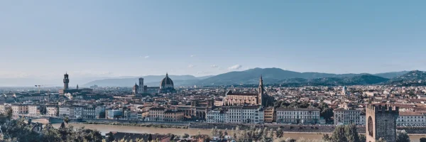 Panoramautsikt over Firenze med elv i forgrunnen, domkirken med rød kuppel og åser i bakgrunnen under klar himmel.