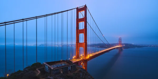 Golden Gate Bridge i skumring med oransje lys langs broen over blått vann og tåke i bakgrunnen.